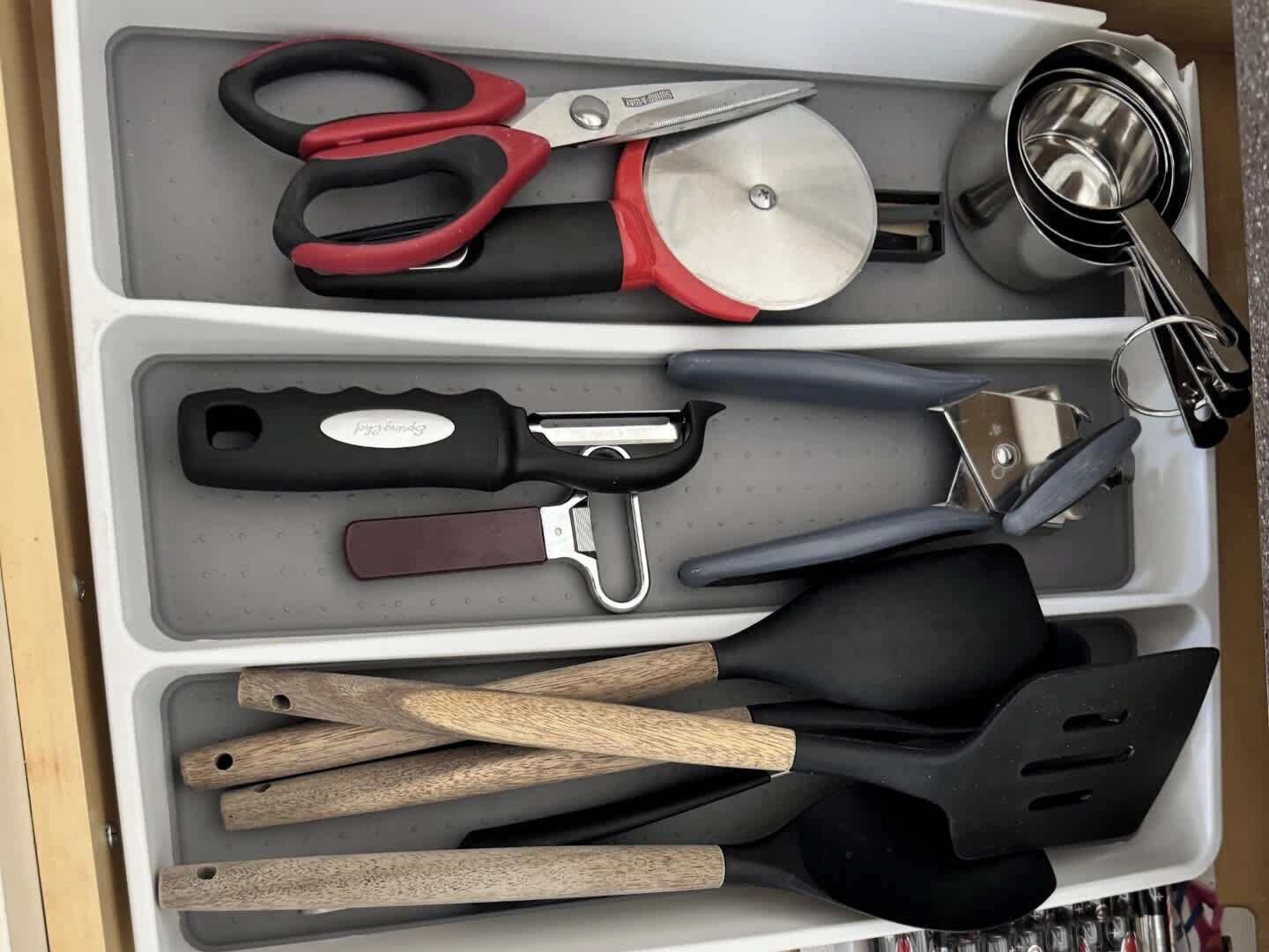 Uma gaveta com diversos utensílios de cozinha, incluindo tesouras, um cortador de pizza, um ralador e espátulas de madeira.