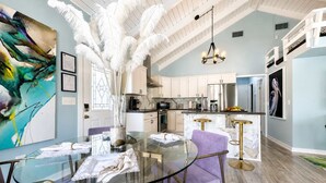 Una cocina moderna con armarios blancos, un refrigerador de acero inoxidable y un comedor con mesa de vidrio y sillas moradas.