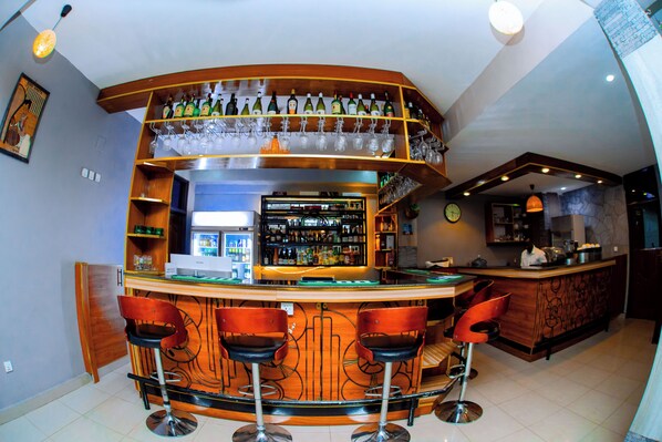 A bar area with a curved counter, multiple stools, shelves with bottles and glasses, and a kitchen area in the background.