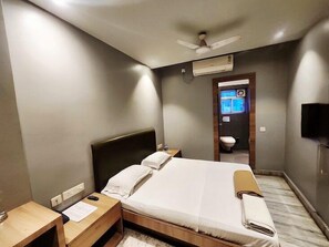 Una habitación de hotel con cama, mesitas de noche, ventilador de techo, unidad de aire acondicionado, televisor y baño visible a través de una puerta abierta.