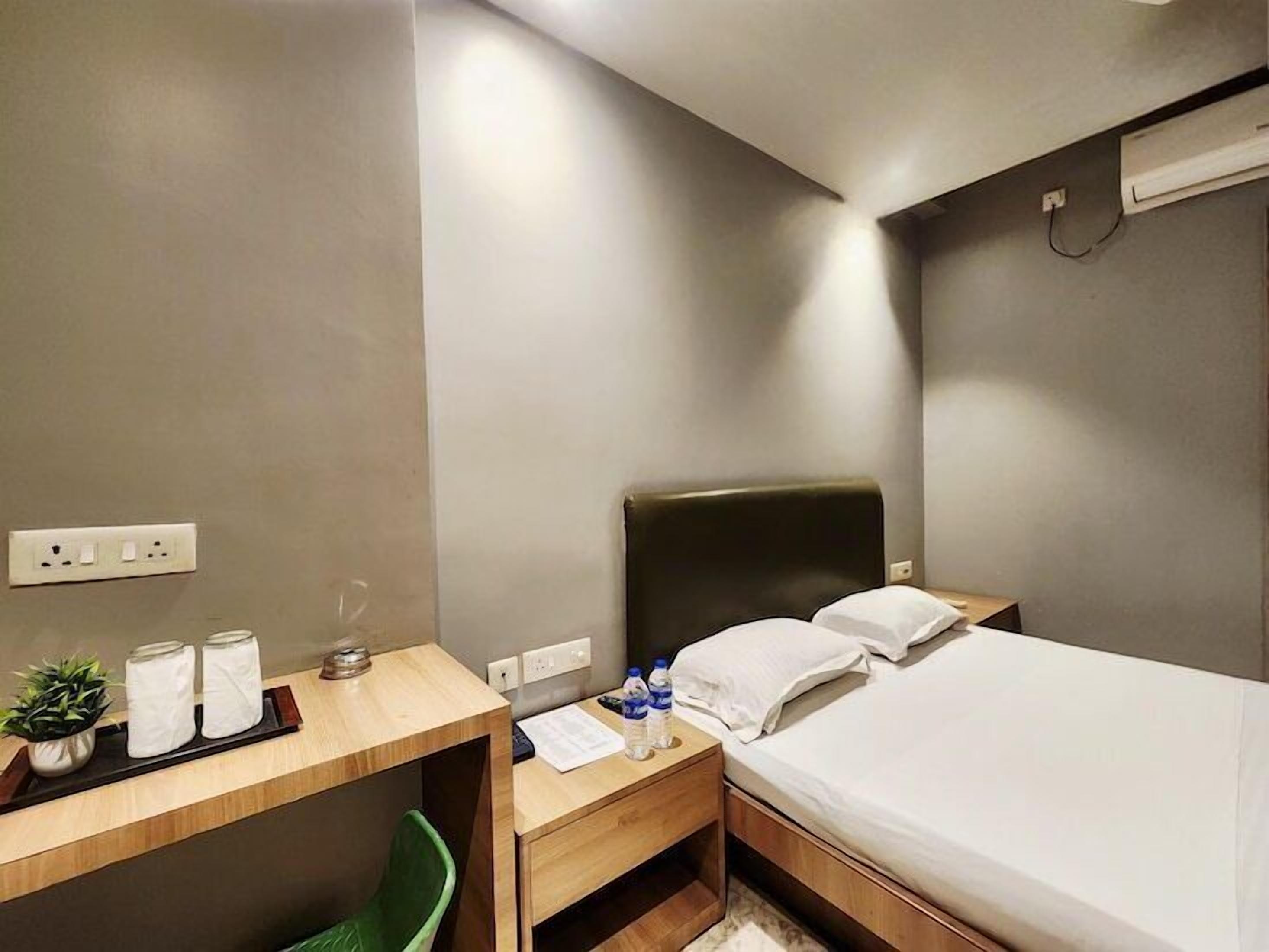 A hotel room with a bed, a desk, a chair, a trash bin, and a small table with toiletries.
