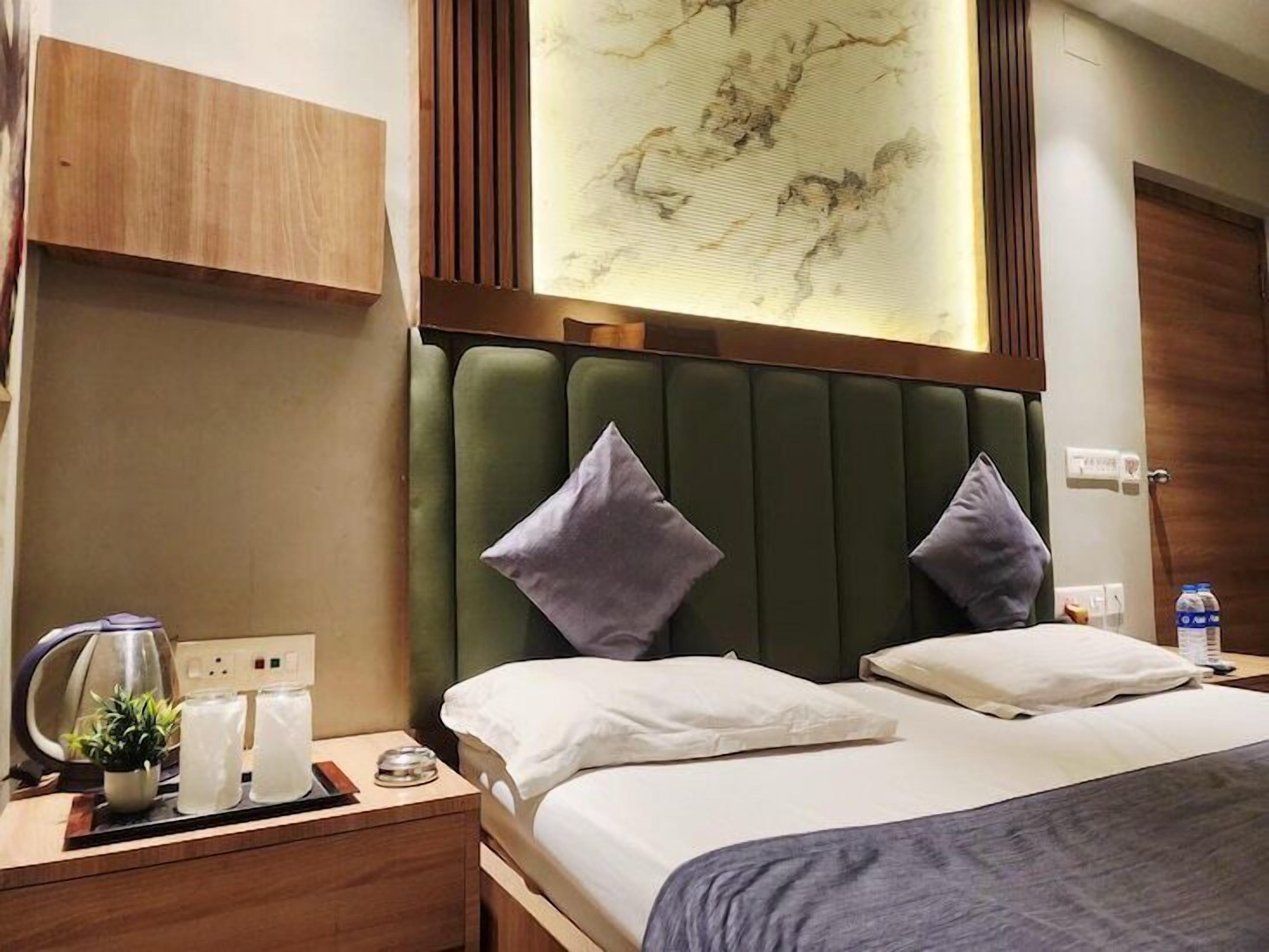A hotel room with a bed, two pillows, a nightstand with a kettle, glasses, and a plant.