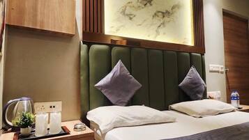 A hotel room with a bed, two pillows, a nightstand with a kettle, glasses, and a plant.