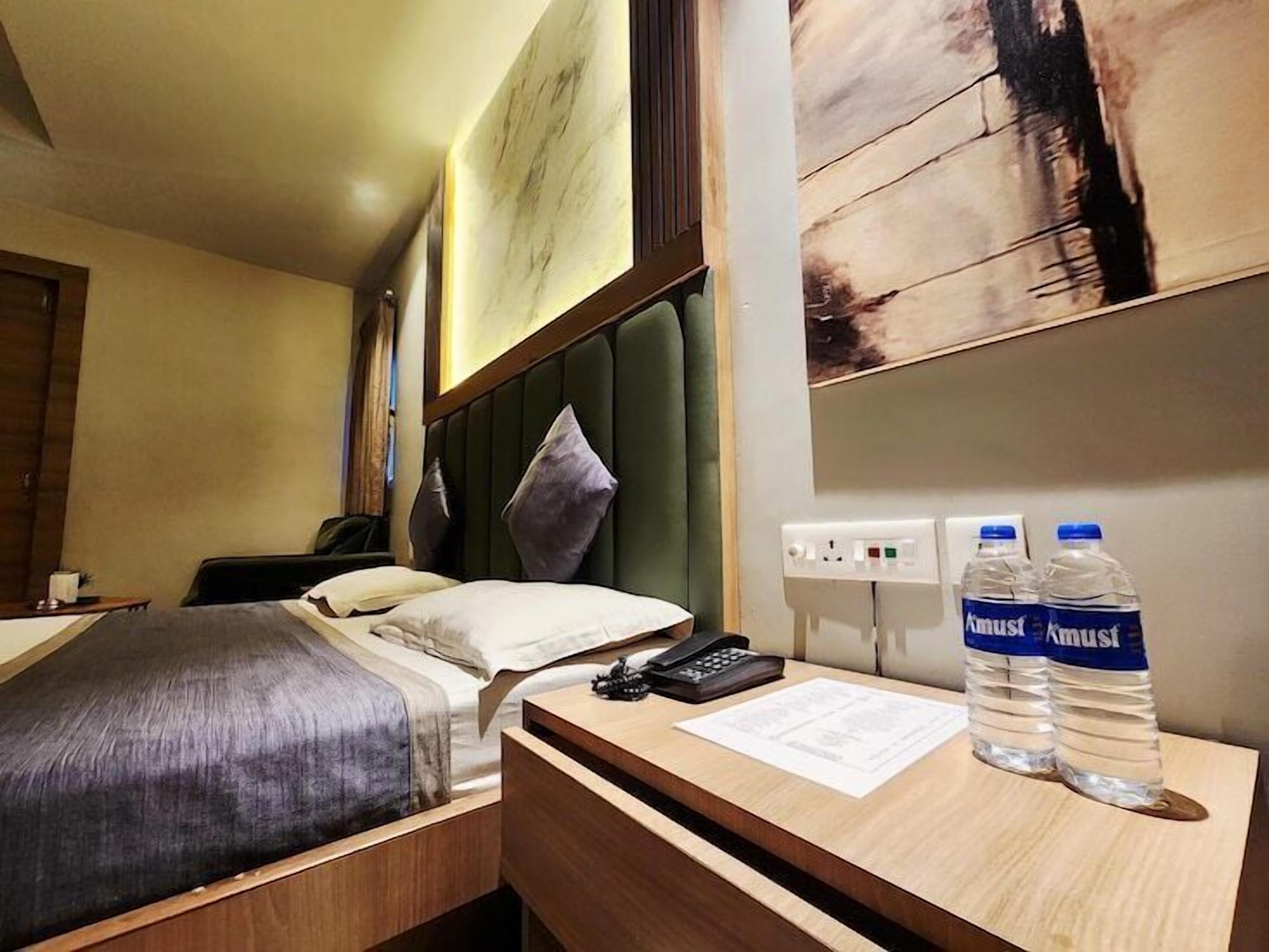 A hotel room with a bed, bedside table, and a desk with two water bottles.