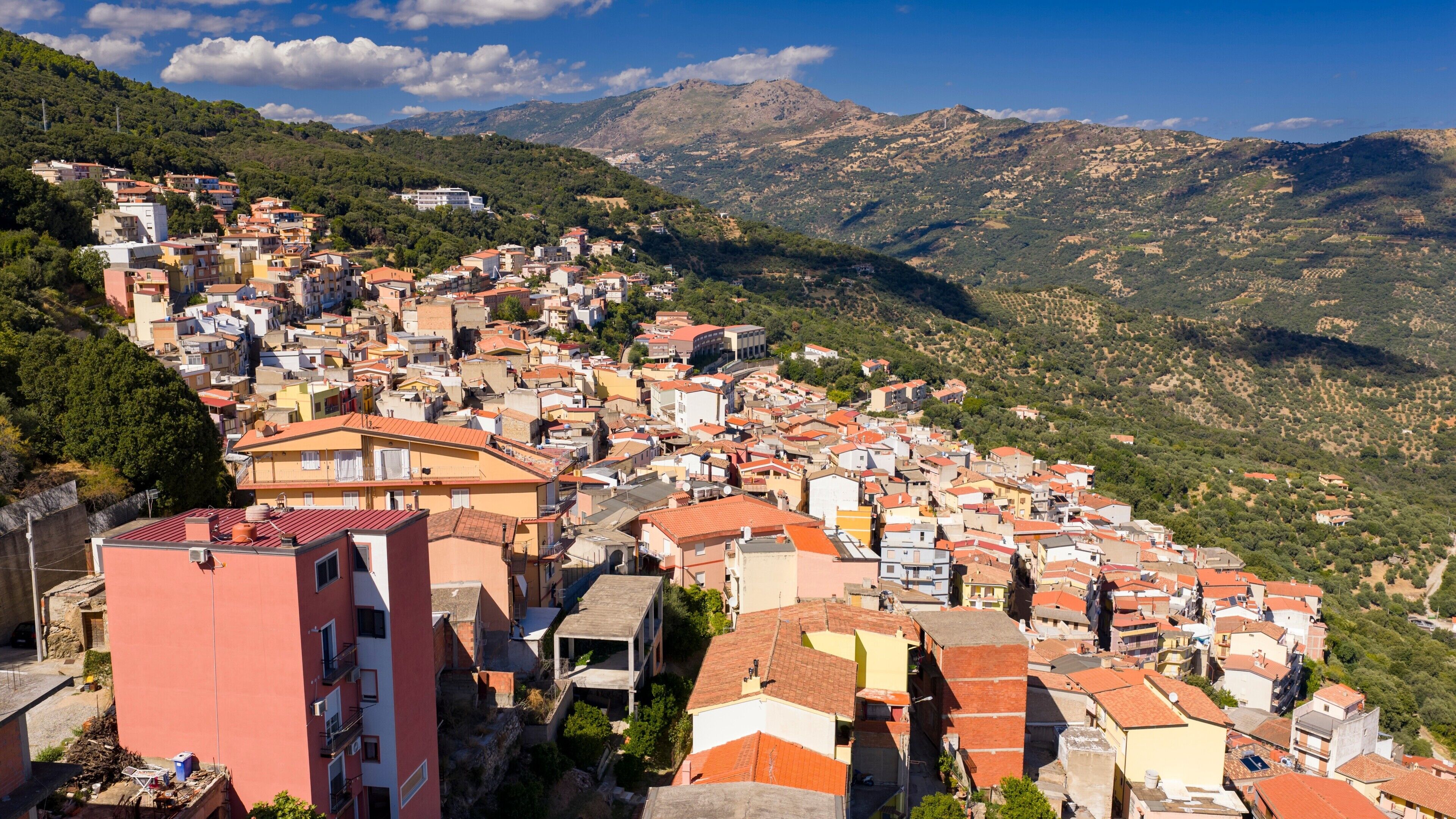 A hillside town with closely packed buildings, a prominent pink building in the foreground, and a mountainous backdrop.