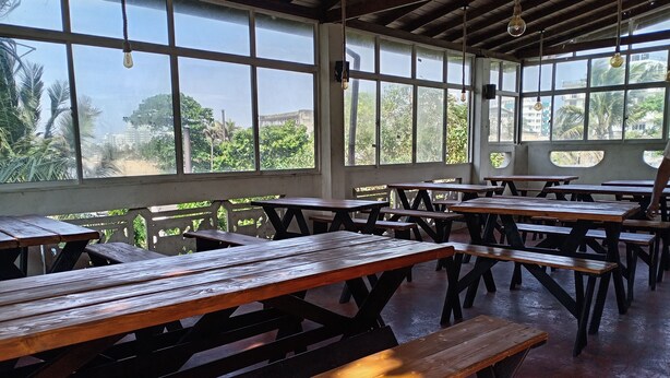 Un espace intérieur vaste et lumineux, avec de nombreuses tables et bancs en bois, de grandes fenêtres laissant entrer la lumière naturelle et une vue sur la verdure environnante.