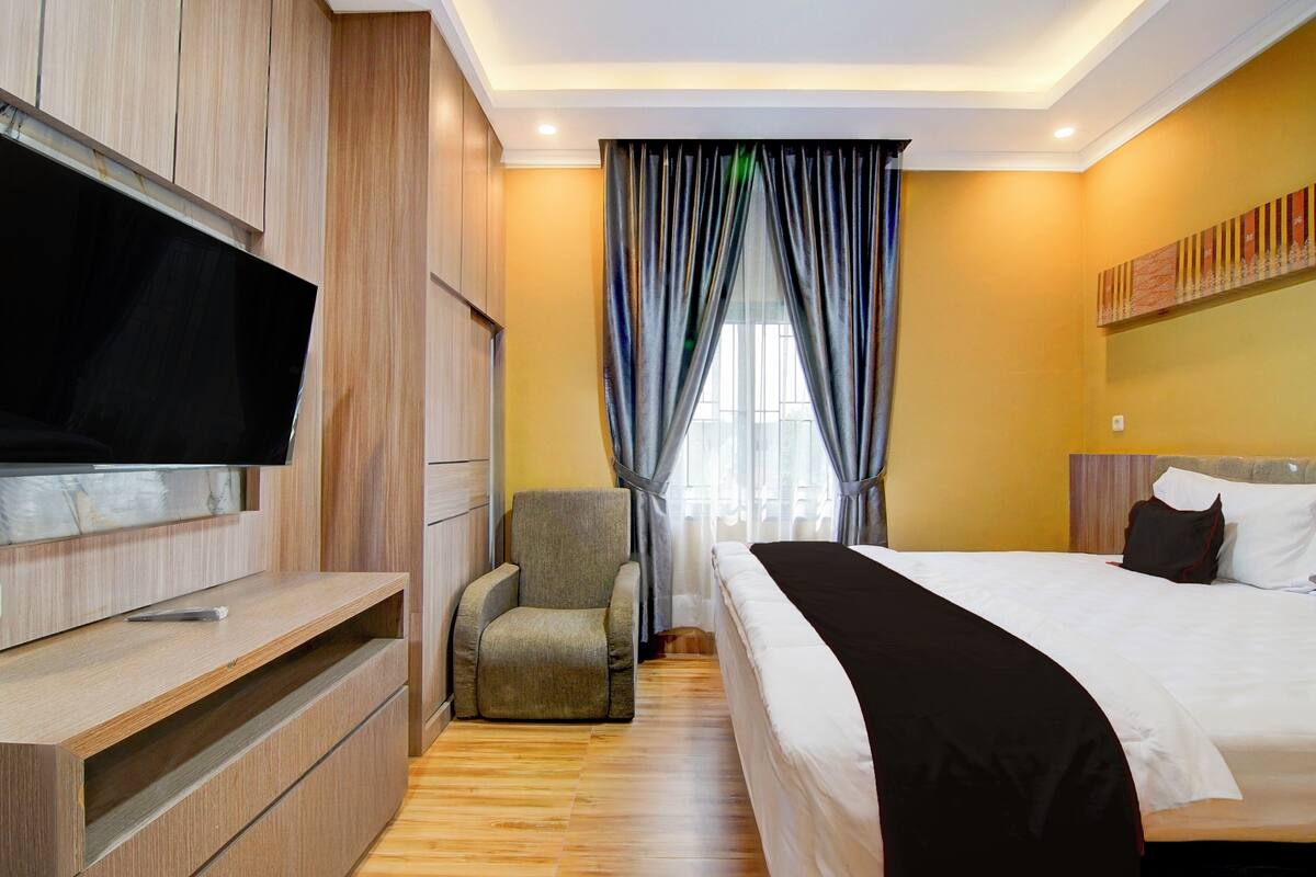 Habitación de hotel con cama, televisor montado en un mueble de madera, una silla y ventana con cortinas.