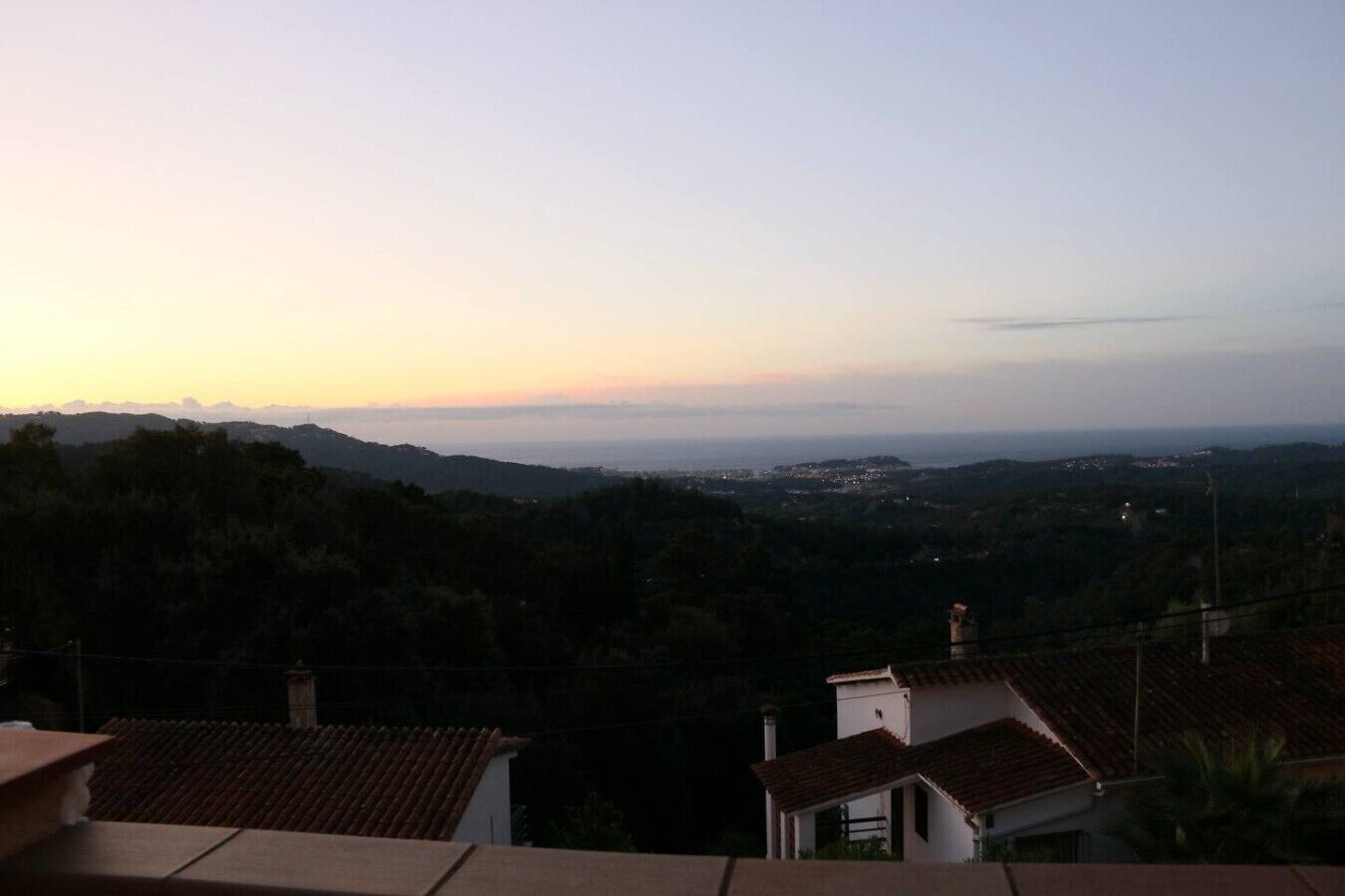 A view of a hilly landscape with a distant town, surrounded by trees and residential buildings with red-tiled roofs.