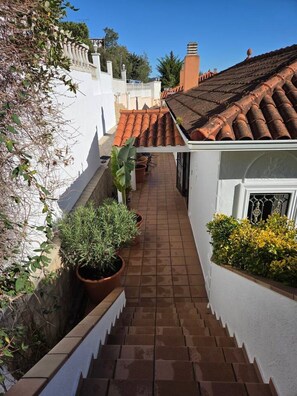 Un estrecho pasillo exterior con suelo de baldosas, plantas en macetas y un edificio blanco con tejado de tejas.
