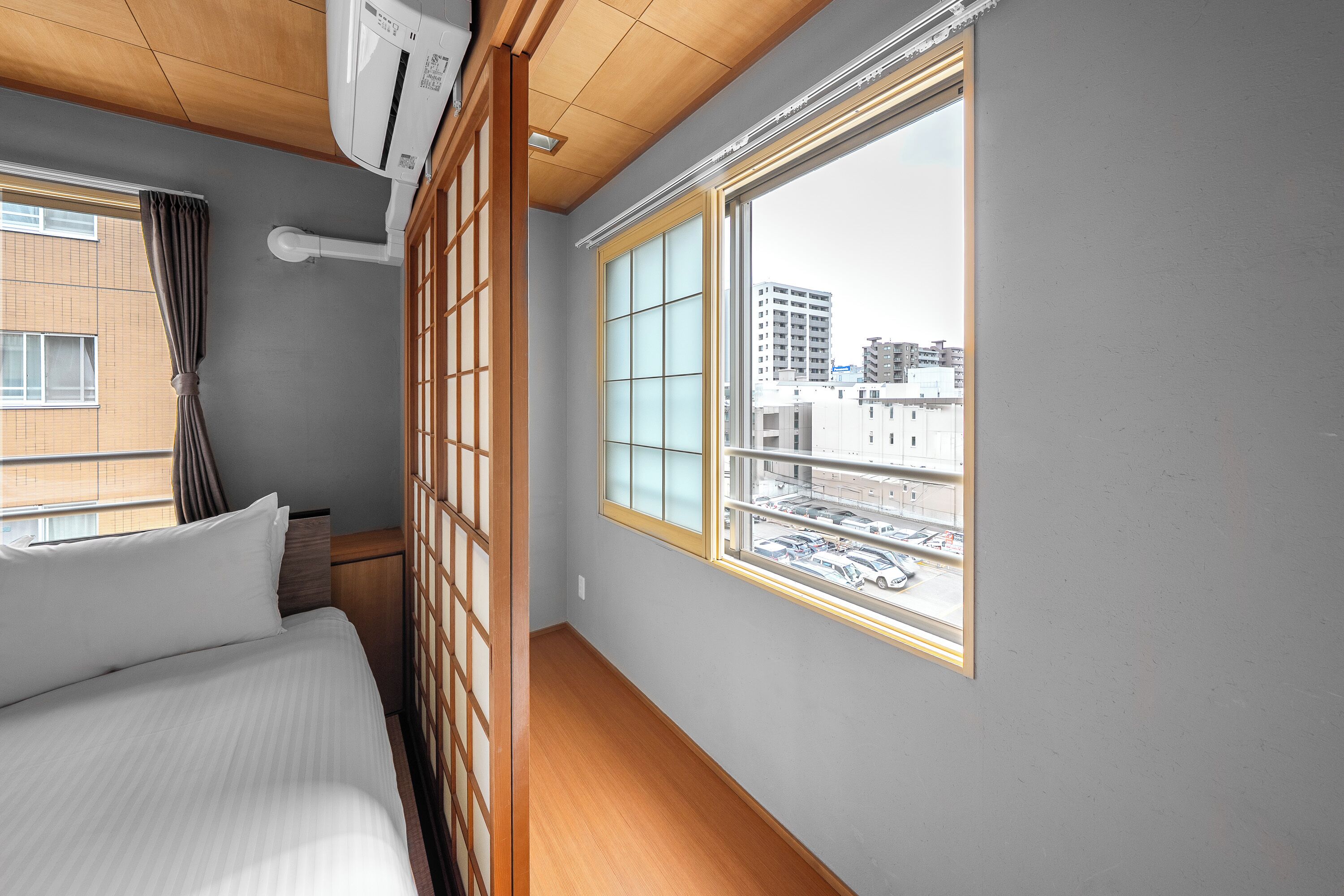 A hotel room with a bed, a window with a view of buildings, and a wooden ceiling.
