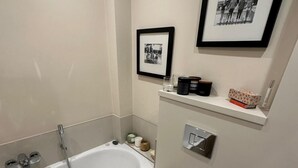 Un baño con bañera, inodoro, lavamanos, repisas con artículos de aseo y cuadros en la pared.