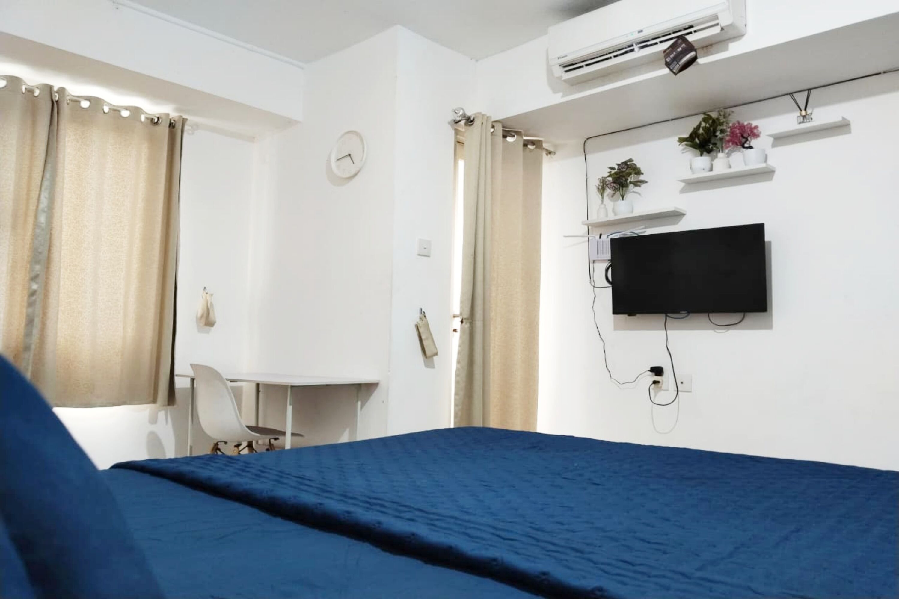 A hotel room with a bed, a desk, a chair, a TV mounted on the wall, and a window with curtains.