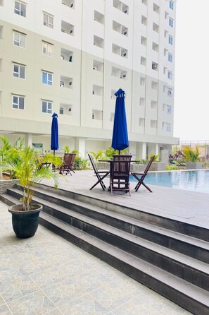 Un espace aménagé au bord de la piscine, avec des marches menant à celle-ci, le tout entouré d’un immeuble à plusieurs étages doté de balcons.