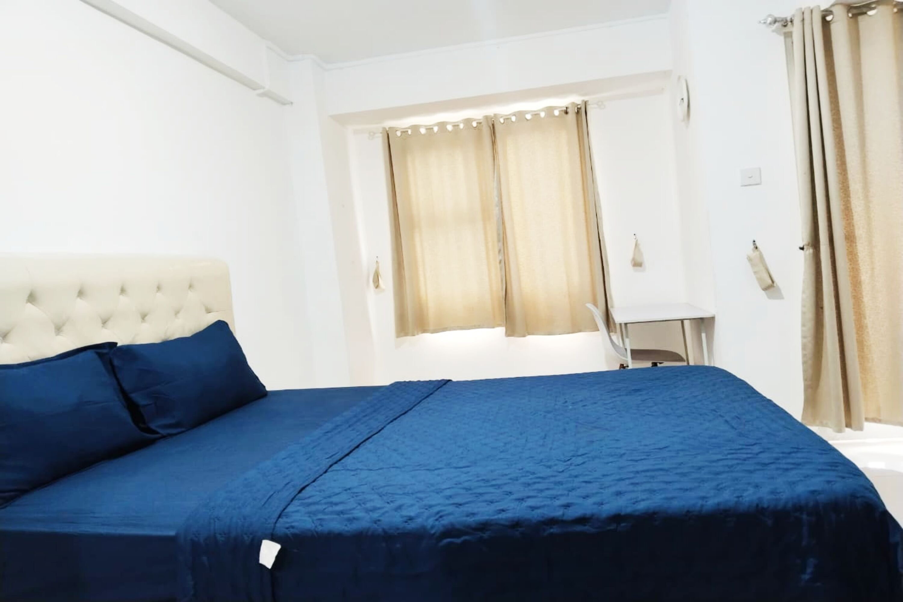 Un letto ben rifatto con copriletto e cuscini blu, una testiera capitonata e una finestra con tende.