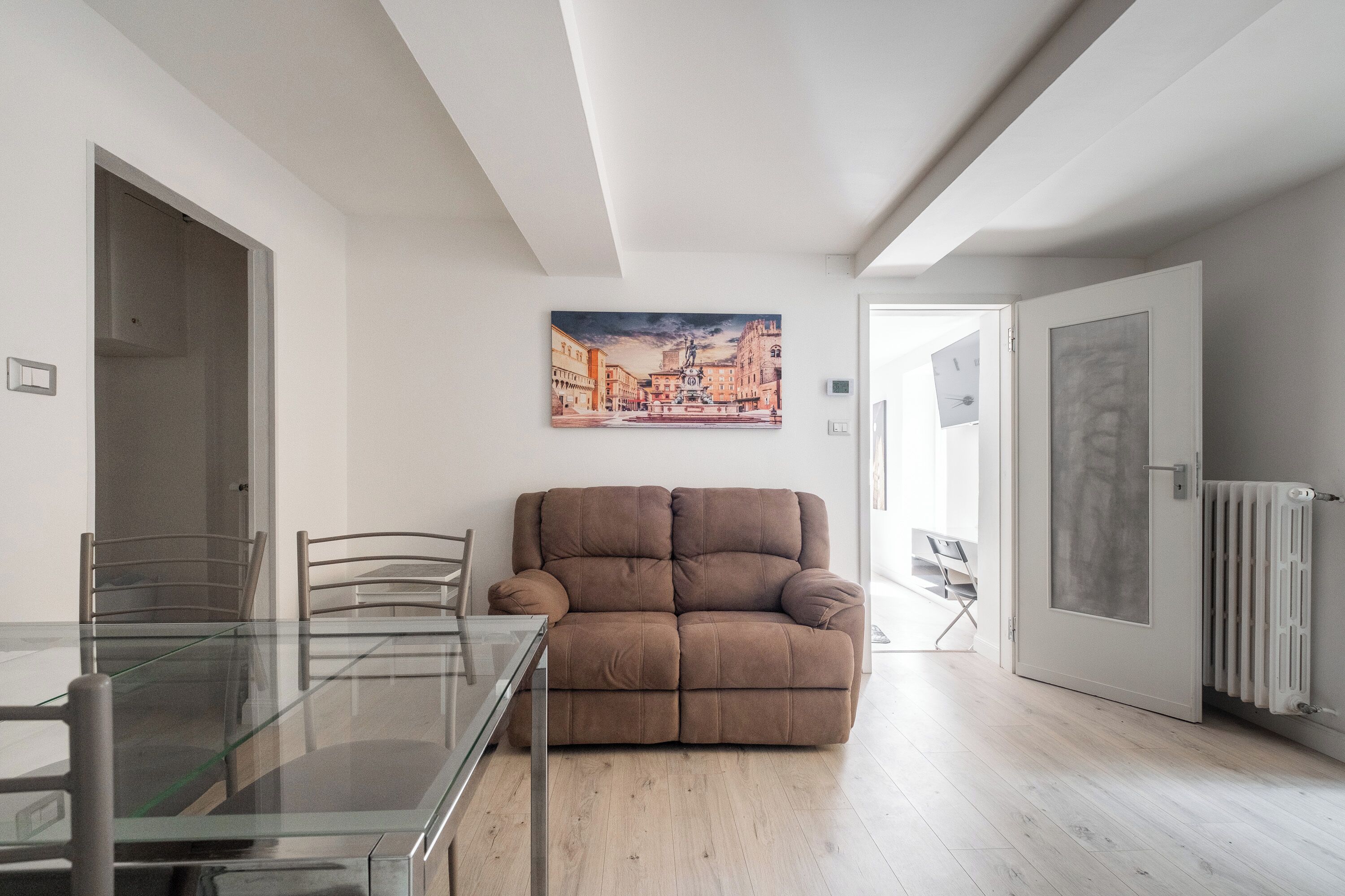 A modern living room with a glass-top dining table, a brown sofa, and a cityscape painting on the wall.