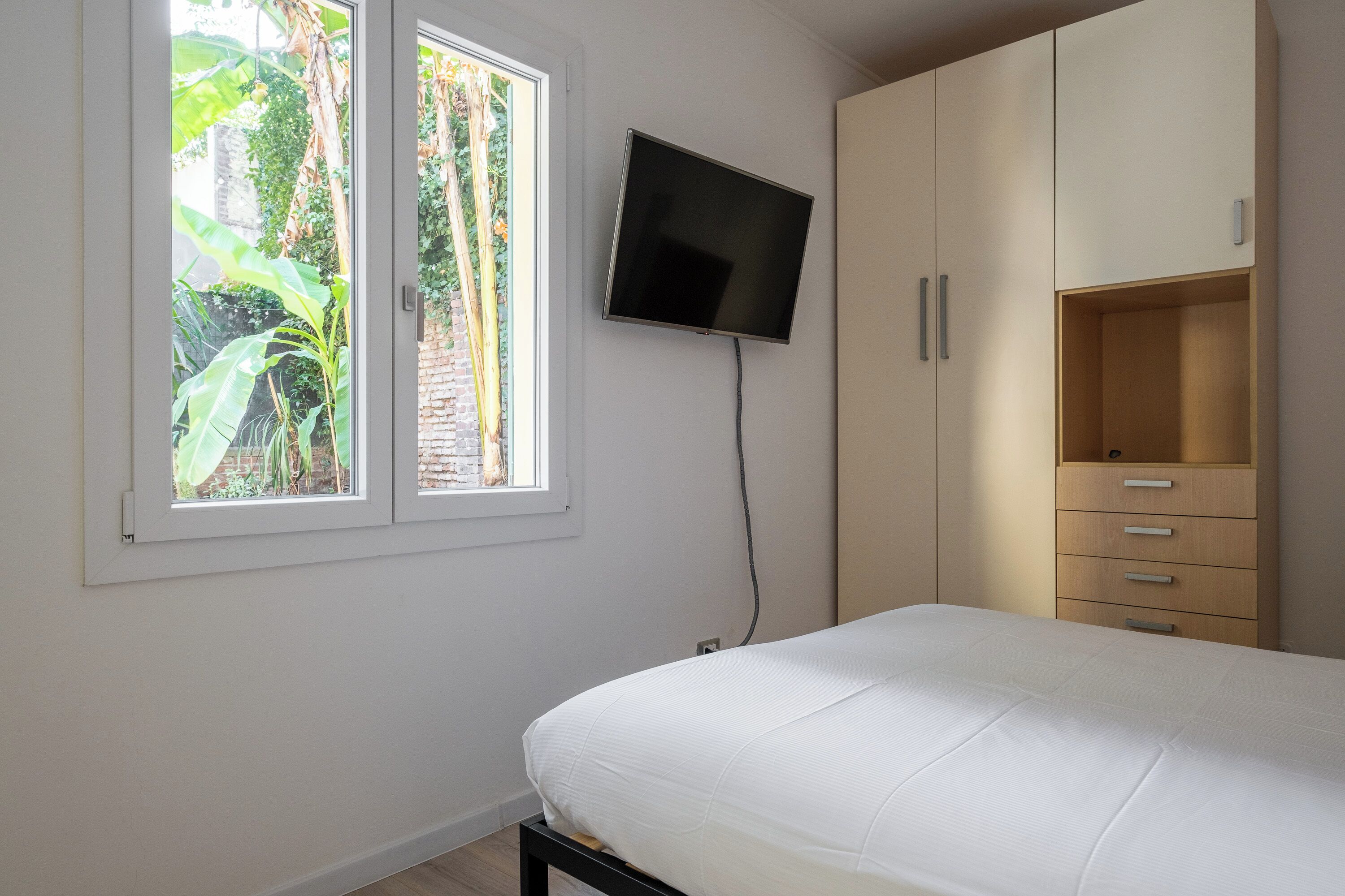 A bedroom with a bed, a TV mounted on the wall, and a window with a view of greenery.