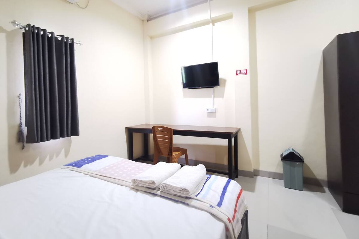 A hotel room with a bed, a desk, a chair, a TV, and a trash bin.