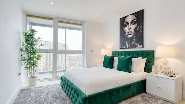 Ein Schlafzimmer mit einem mit grünem Stoff bezogenen Bett, einem großen Fenster, einer Topfpflanze und einem Gemälde an der Wand.
