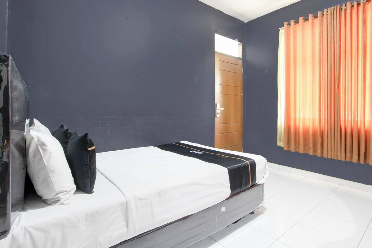 A bedroom with a bed, pillows, a suitcase, a window with curtains, and a door.