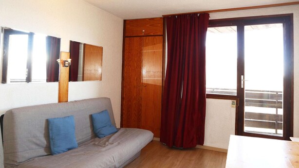 Una habitación con un sofá gris, un armario de madera y una puerta corredera de vidrio con cortinas rojas.