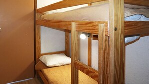 Una litera con estructura de madera, una sola almohada y un pequeño estante con una lámpara.