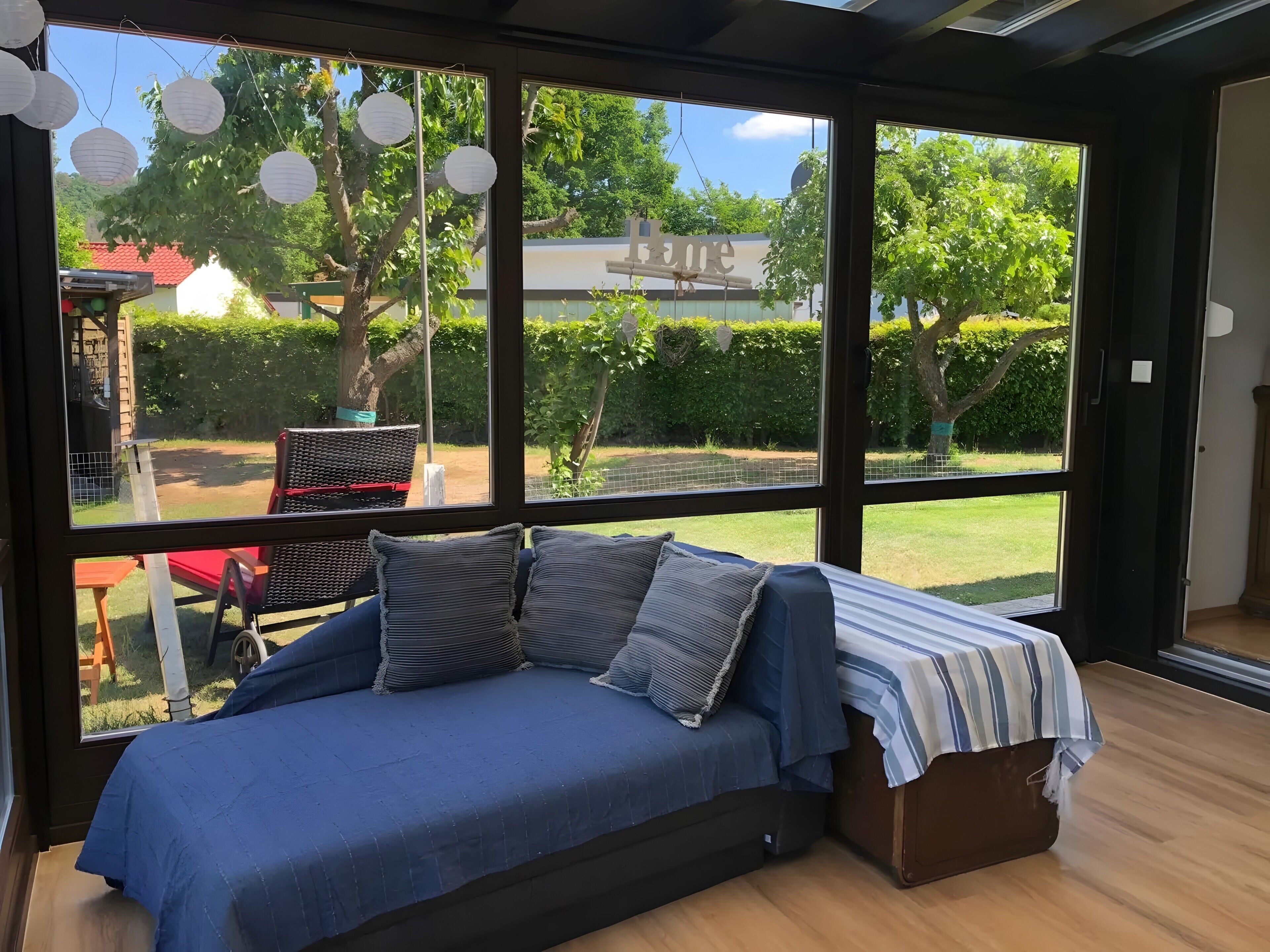 A room with a large window, a bed with a blue bedspread, and a view of a garden and trees.