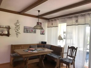 Un coin repas avec une table et un banc en bois, un luminaire suspendu et une vue dégagée à travers des rideaux légers.