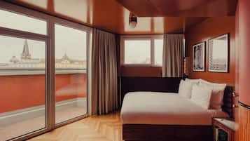 Habitación de hotel con una cama grande, una mesita de noche, una estantería y grandes ventanas con vistas a la ciudad.