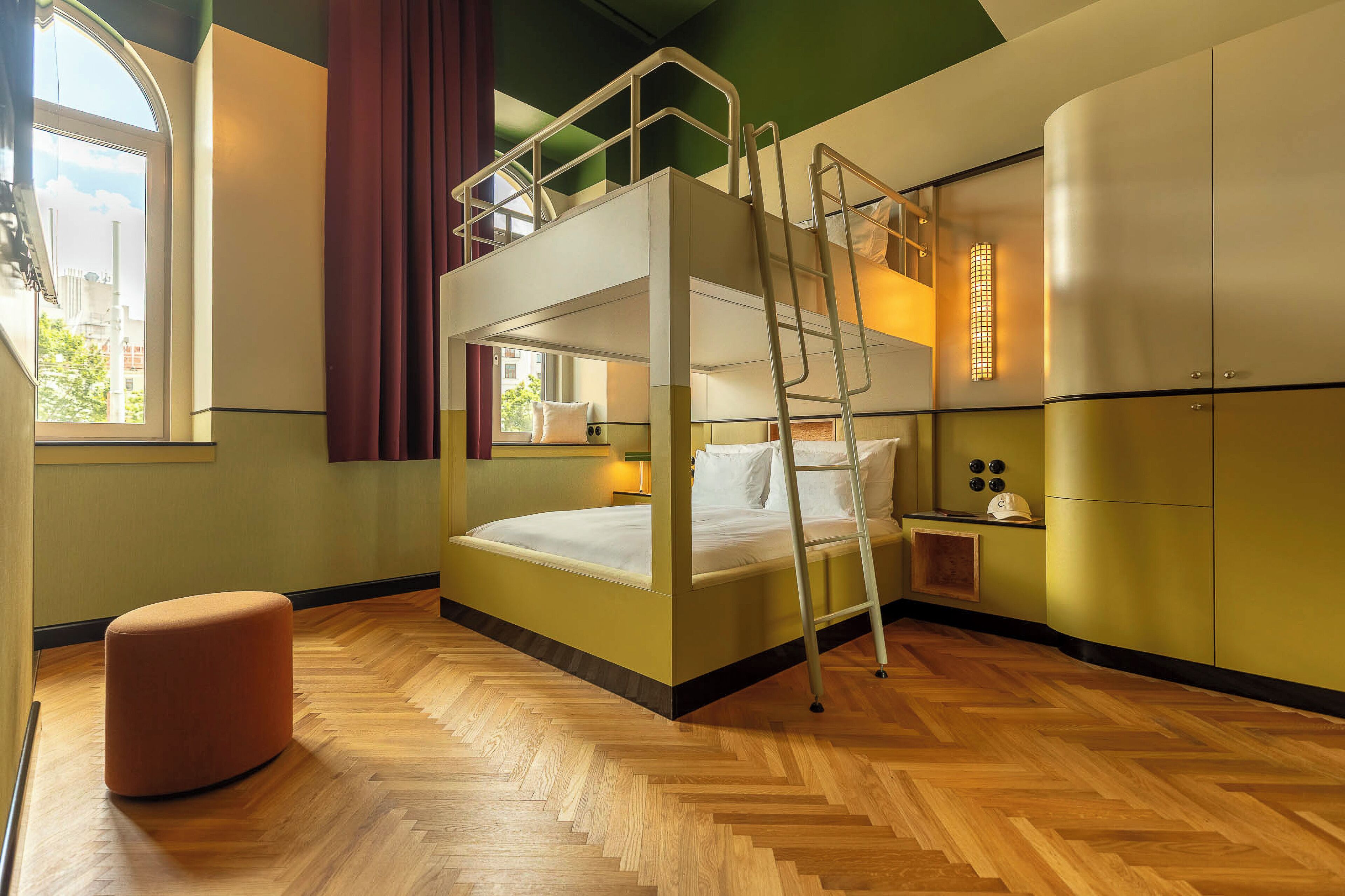 A bunk bed room with a wooden floor, a desk with a lamp, and a window with curtains.