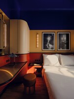 Quarto de hotel com uma cama, uma escrivaninha, uma cadeira, uma mesinha de cabeceira com uma luminária e duas imagens emolduradas na parede.