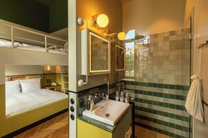 Quarto de hotel com uma cama beliche, banheiro com um espelho grande, pia e chuveiro.