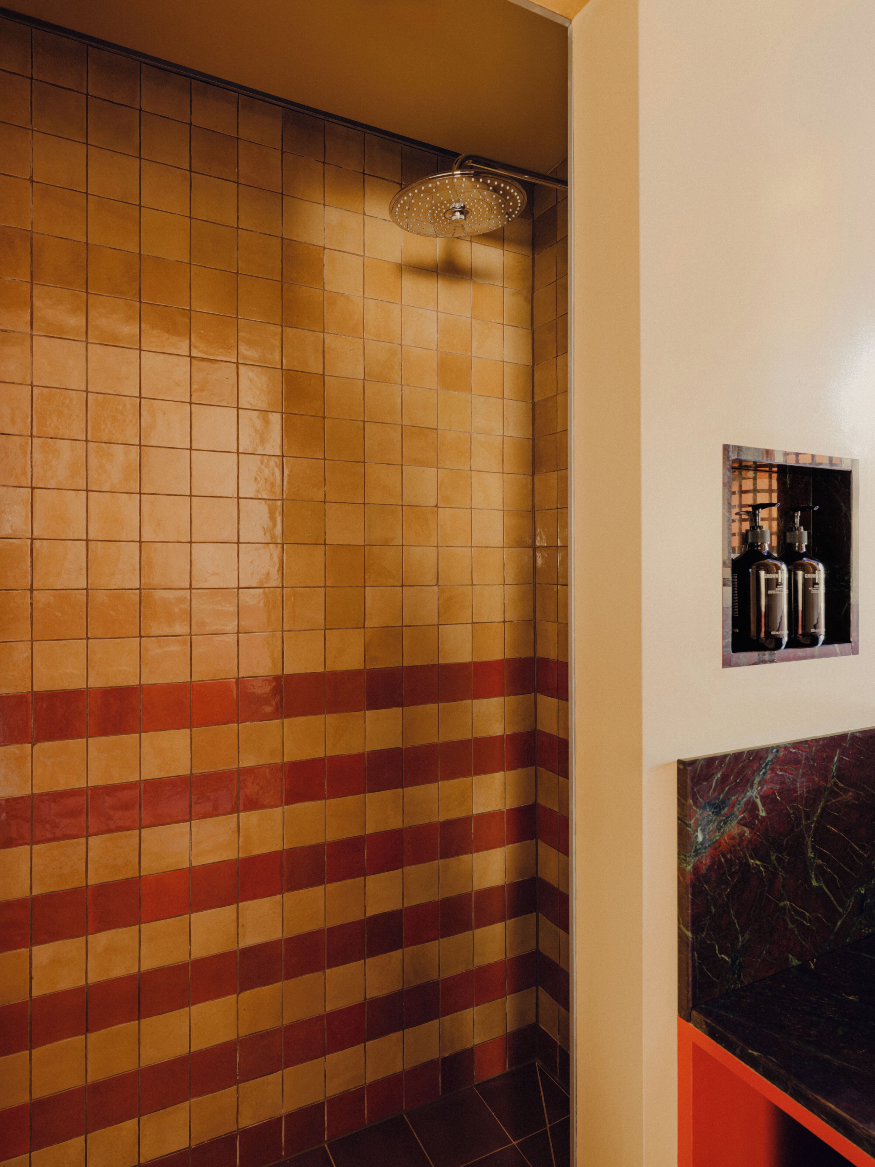 Un baño con paredes de azulejos, ducha y repisa con botellas.