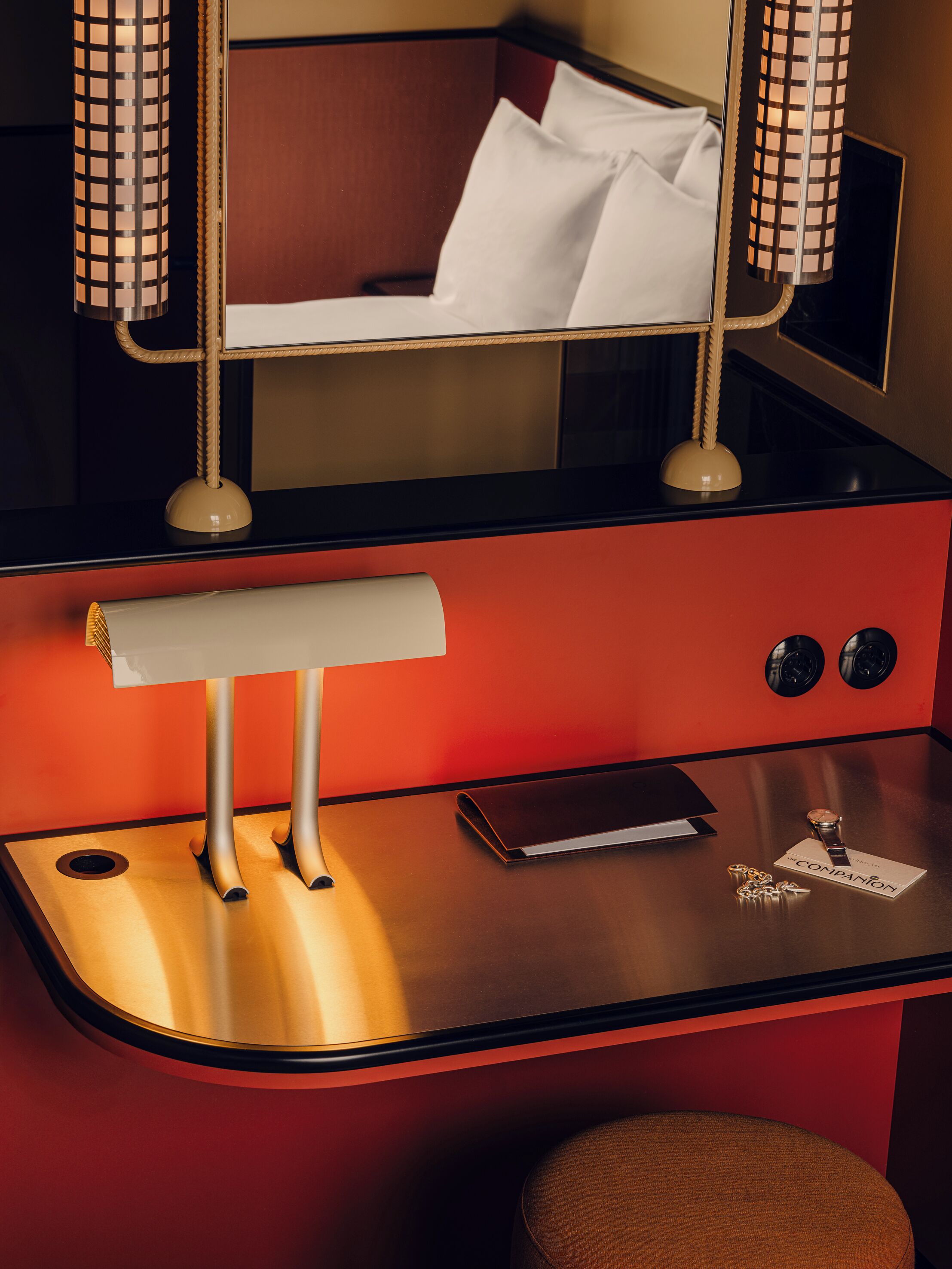 Une chambre d’hôtel compacte comprenant un lit superposé, un bureau avec une lampe, une petite table, une chaise et une table de chevet avec une montre et des clés.