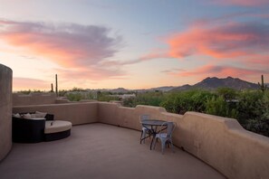 Une terrasse sur le toit, avec une table ronde et des chaises, un canapé, et une vue sur les montagnes et la végétation, le tout sous un ciel aux couleurs vives.