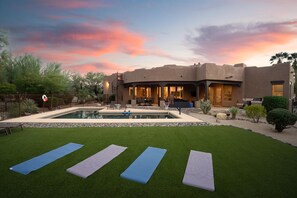 Une maison moderne avec une piscine, des tapis de yoga sur la pelouse et un ciel au coucher du soleil.