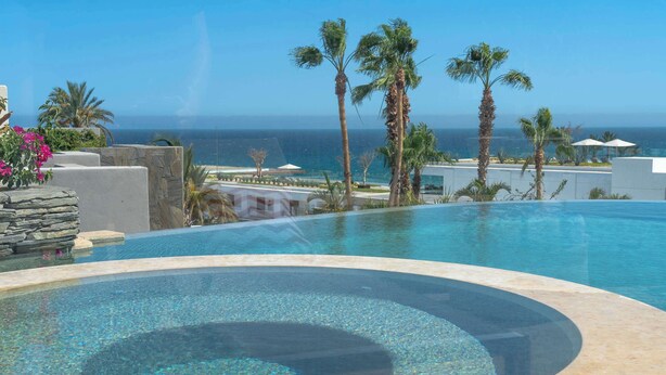 A clear blue infinity pool with a pebble bottom, surrounded by palm trees and a view of the ocean. - 6BR Luxe Villa w/ Infinity Pool & Ocean Views (Cabo San Lucas)