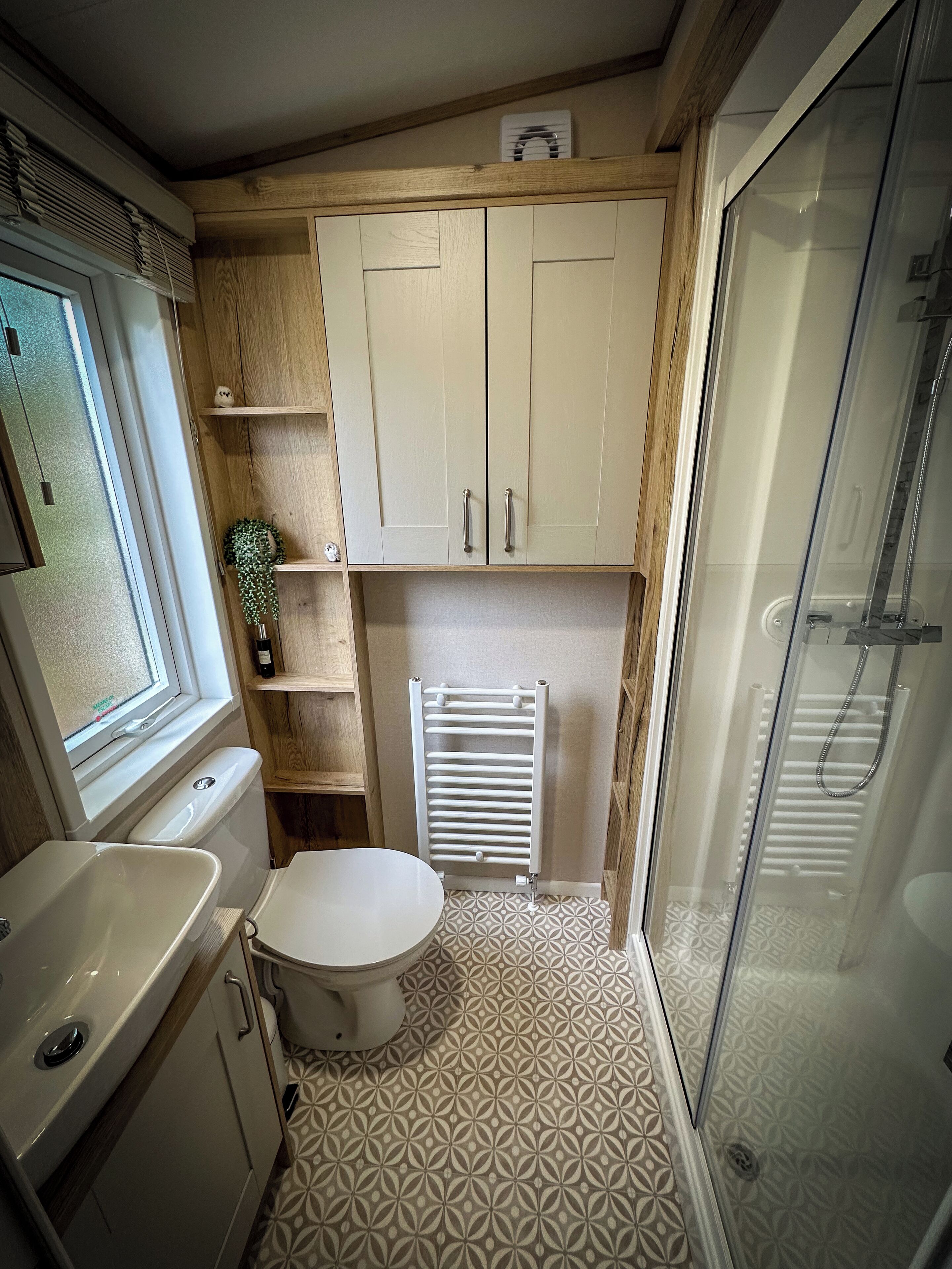 A compact bathroom with a toilet, sink, and shower. It features wooden cabinetry, a radiator, and patterned floor tiles.