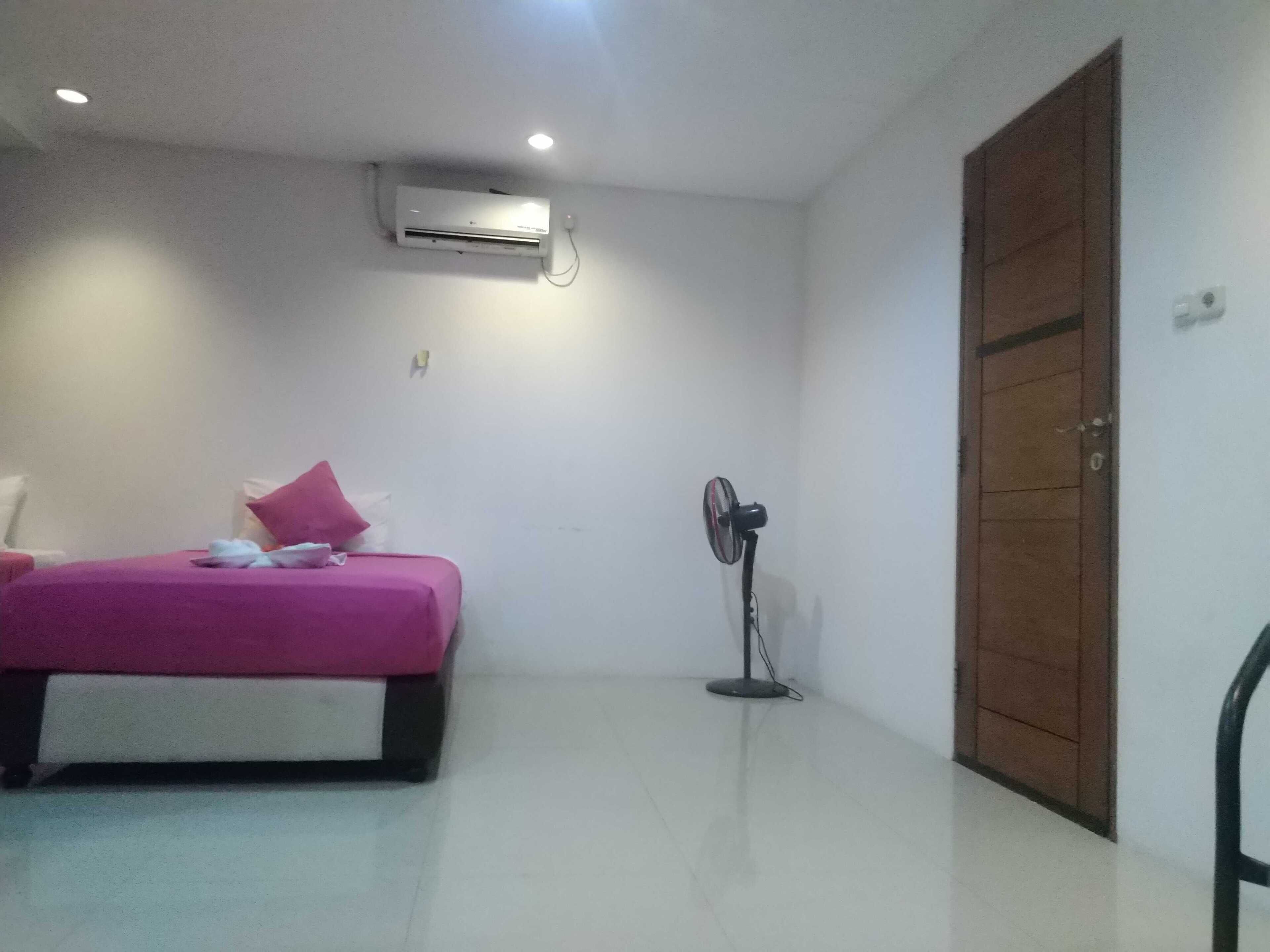 A bedroom with a bed, an air conditioner, a standing fan, and a wooden wardrobe.