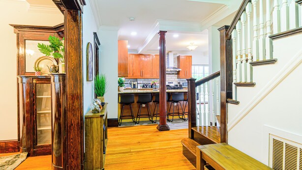 A wooden-floored hallway leading to a kitchen with bar stools and a staircase with white balusters.