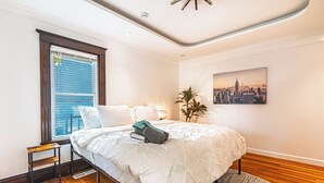 A bedroom with a bed, a window with blinds, a small wooden nightstand, a cityscape painting on the wall, and a plant.