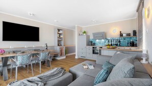 A modern living room with a grey sofa, clear chairs, a flat-screen TV, and a kitchen area with a blue backsplash.