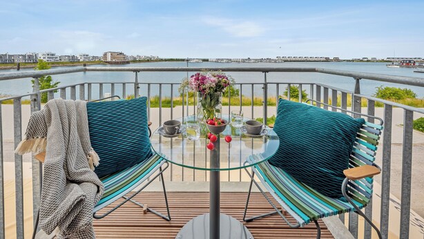 A glass-top table with two chairs, a bowl of fruit, and a vase of flowers on a balcony overlooking a body of water.