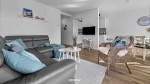 A modern living room with a grey sofa, a wicker chair, a small white table with a plant, and a mounted clock. - Küstenglück - Shower room, covered balcony, garden furniture, TV, parking space (Damp)