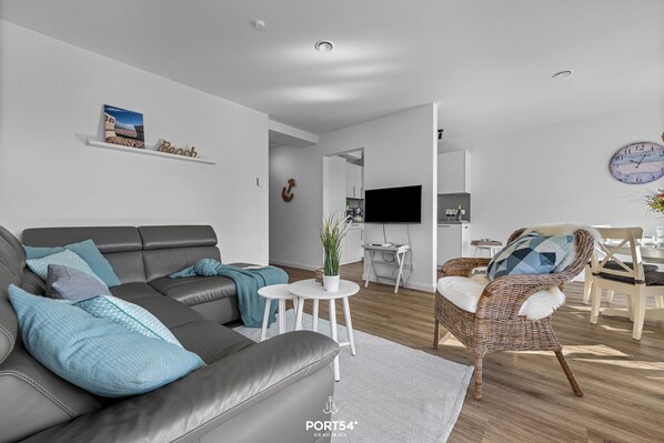 A modern living room with a grey sofa, a wicker chair, a small white table with a plant, and a mounted clock. - Küstenglück - Shower room, covered balcony, garden furniture, TV, parking space (Damp)