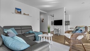 Un salon moderne avec un canapé gris, un fauteuil en osier, une petite table blanche ornée d’une plante et une horloge murale.