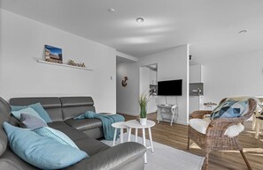 A modern living room with a grey sofa, a wicker chair, a small white table with a plant, and a mounted clock. - Küstenglück - Shower room, covered balcony, garden furniture, TV, parking space (Damp)