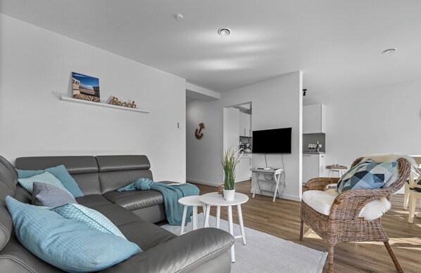 Una sala de estar moderna con un sofá gris, una silla de mimbre, una mesita blanca con una planta y un reloj de pared.