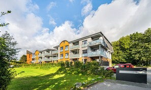 A multi-story residential building with balconies, a grassy area, and a red car parked nearby. - Küstenglück - Shower room, covered balcony, garden furniture, TV, parking space (Damp)