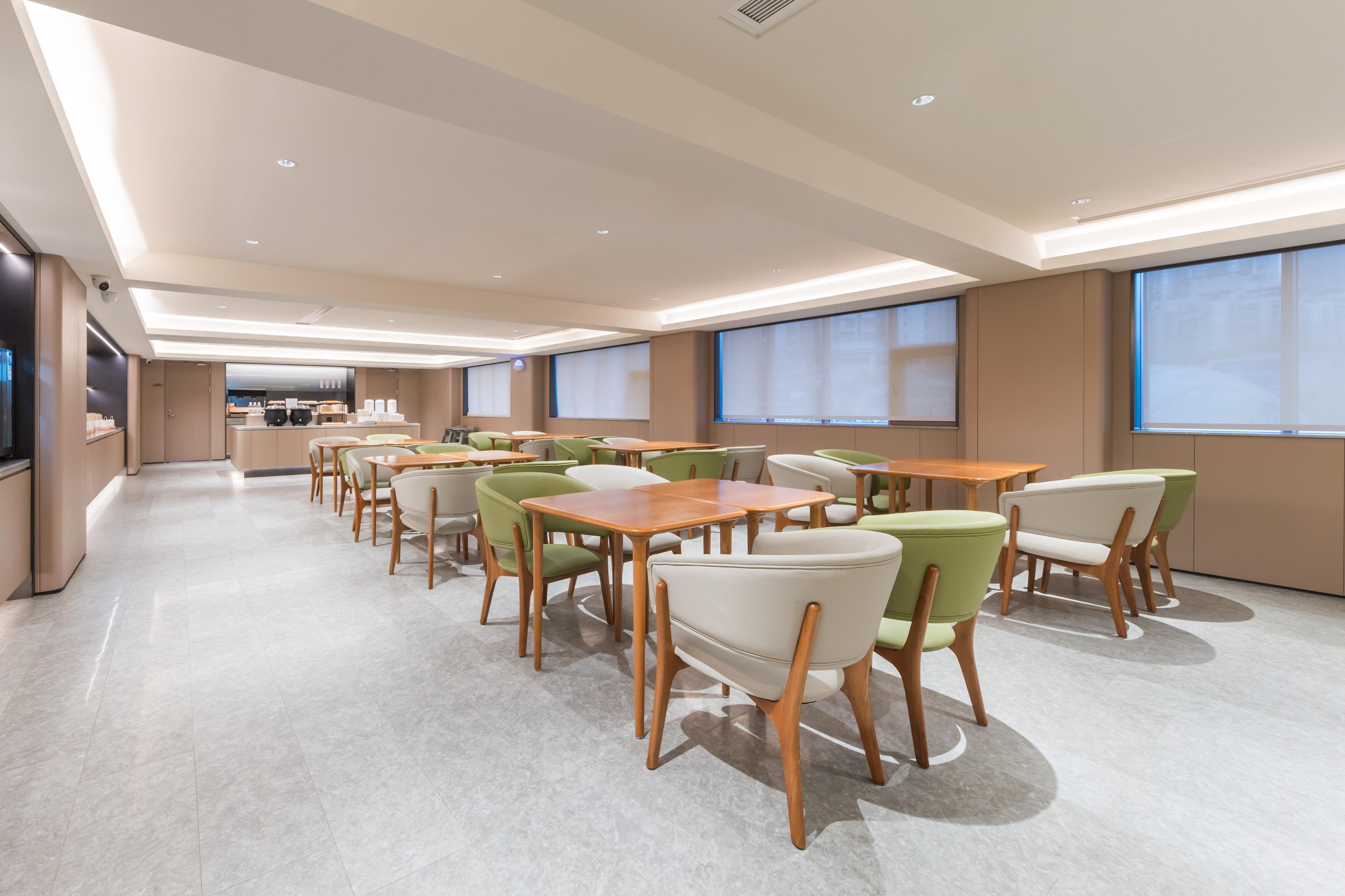 A spacious dining area with multiple tables and chairs, a kitchen in the background, and large windows providing natural light.