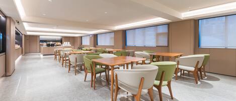 A spacious dining area with multiple tables and chairs, a kitchen in the background, and large windows providing natural light.